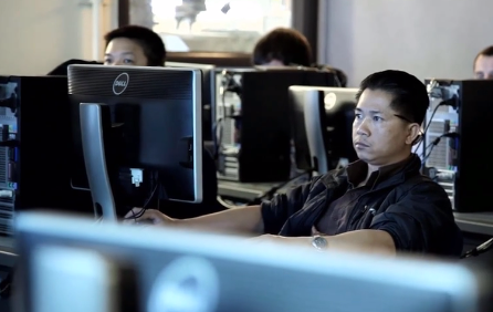 A man in a black shirt in a computer lab with his arms stretched out.