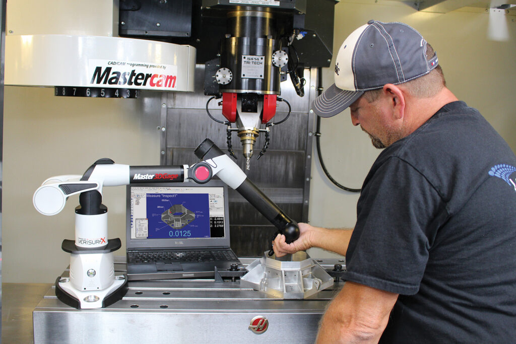 A man in a baseball cap holding the end of the Master3DGage scanner arm on a CNC machine with Verisurf on the laptop screen behind them.. 