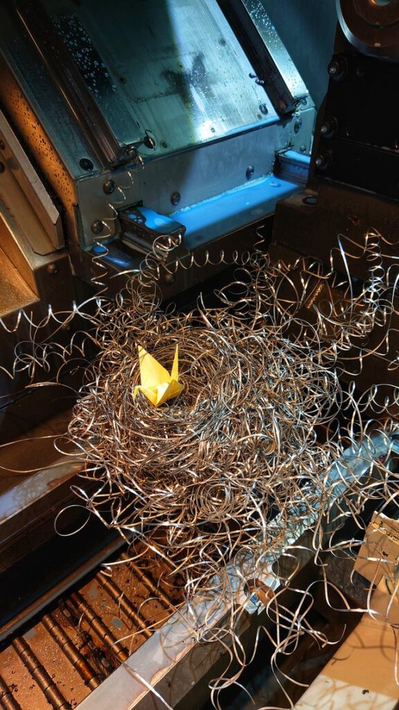 A yellow origami crane on a spiral of long strands of metal shavings under a CNC machine.