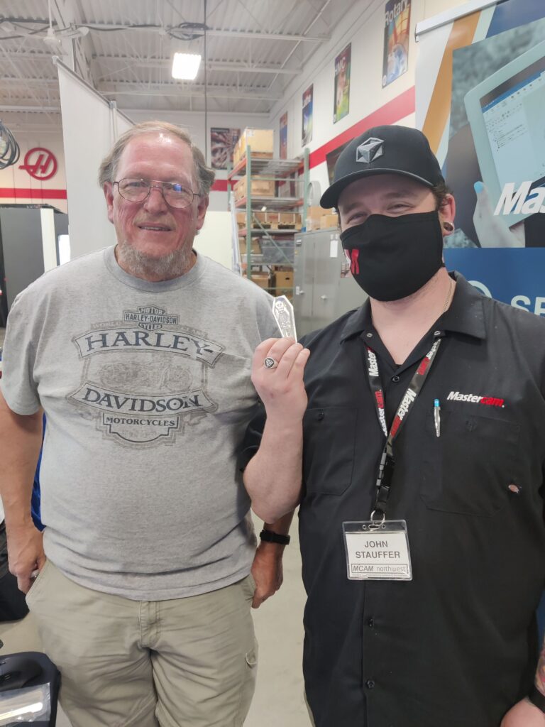 Two mean posing for a picture with Haas logos on the walls. A man with glasses and a Harley Davidson T-shirt (Instructor Jim Kellis) and a man wearing a black Mastercam collared shirt, and black Postability hat (John Stauffer), holding a tiny metal skeleton in a coffin.