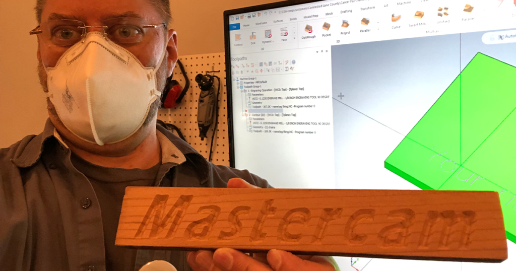 A man with glasses and a mask (Chris Hehman) holding a wooden sign that says "Mastercam" in front of. computer screen with a green 3D model displayed on it.