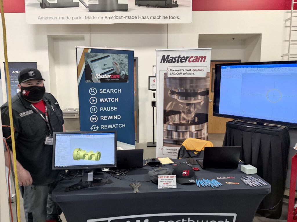 A man with a black mask and black mcam collared shirt (Geoff Phoenix) standing behind a table with computer monitors and banners for Mastercam and Streamingteacher.