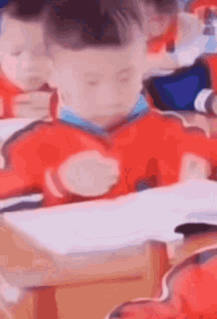 A child in a red tracksuit with blue and white stripes at his desk turning the pages of a book and making a splashing motion with his hands against his face.