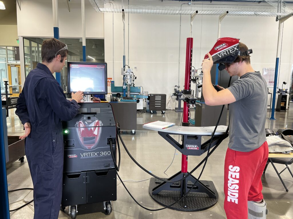 2 young male students using a VRTEX 360 virtual reality helmet on the shop floor of the PCC OMIC Training Center.