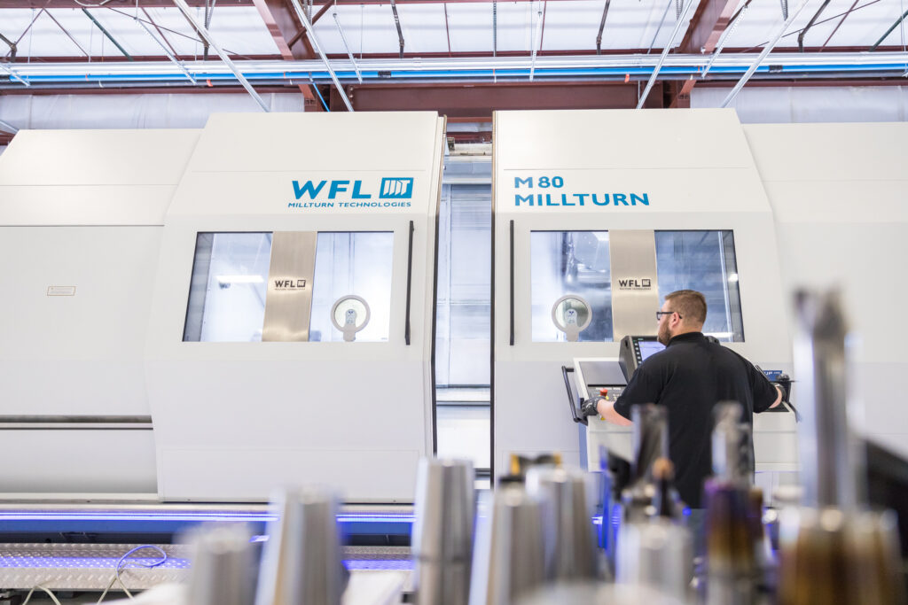 a man with glasses operating an all white WFL Mill-Turn machine with blue lettering with tool holders in the foreground.