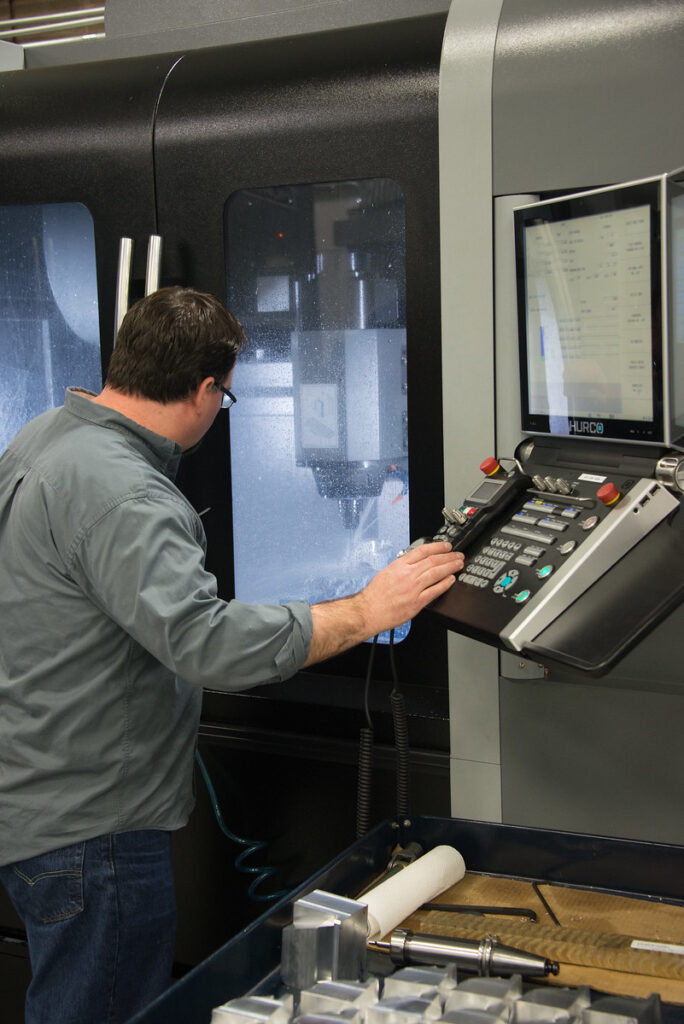 A man in a grey dress shirt and jeans with his hand on the control panel of a CNC machine.