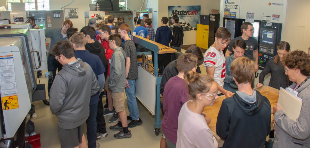A crowd of teenage students in a room looking at CNC machines.